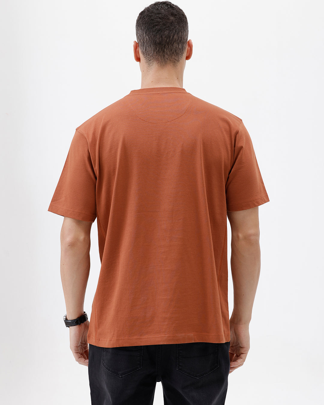 A man wearing a rust-orange crewneck t-shirt with a subtle tonal 'ELECTRIC' logo across the chest, paired with black pants, photographed against a clean white studio background.