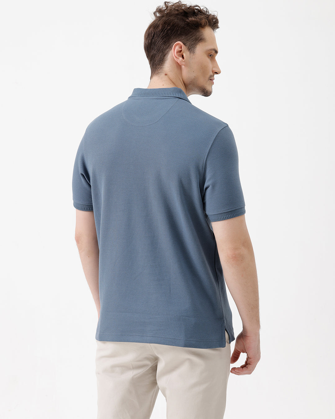 A man wearing a slate blue short-sleeve polo shirt and beige trousers, standing against a plain white studio background.