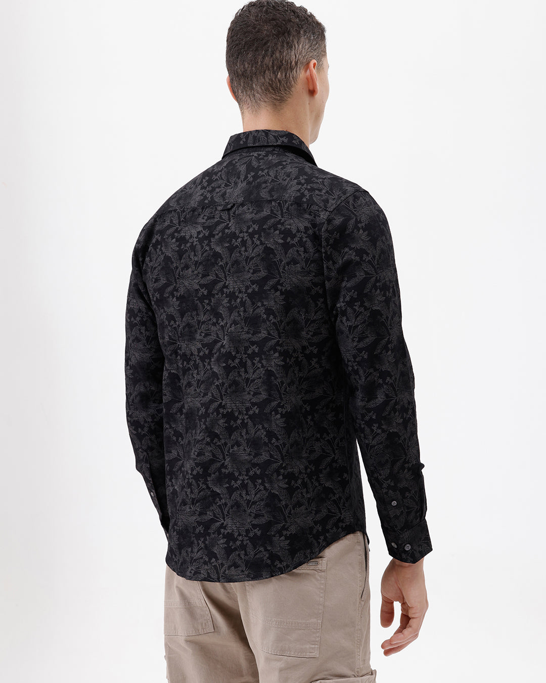 A man wearing a black long-sleeve button-up shirt with a subtle tonal floral pattern, paired with beige cargo pants and dark sunglasses, standing against a plain white background.