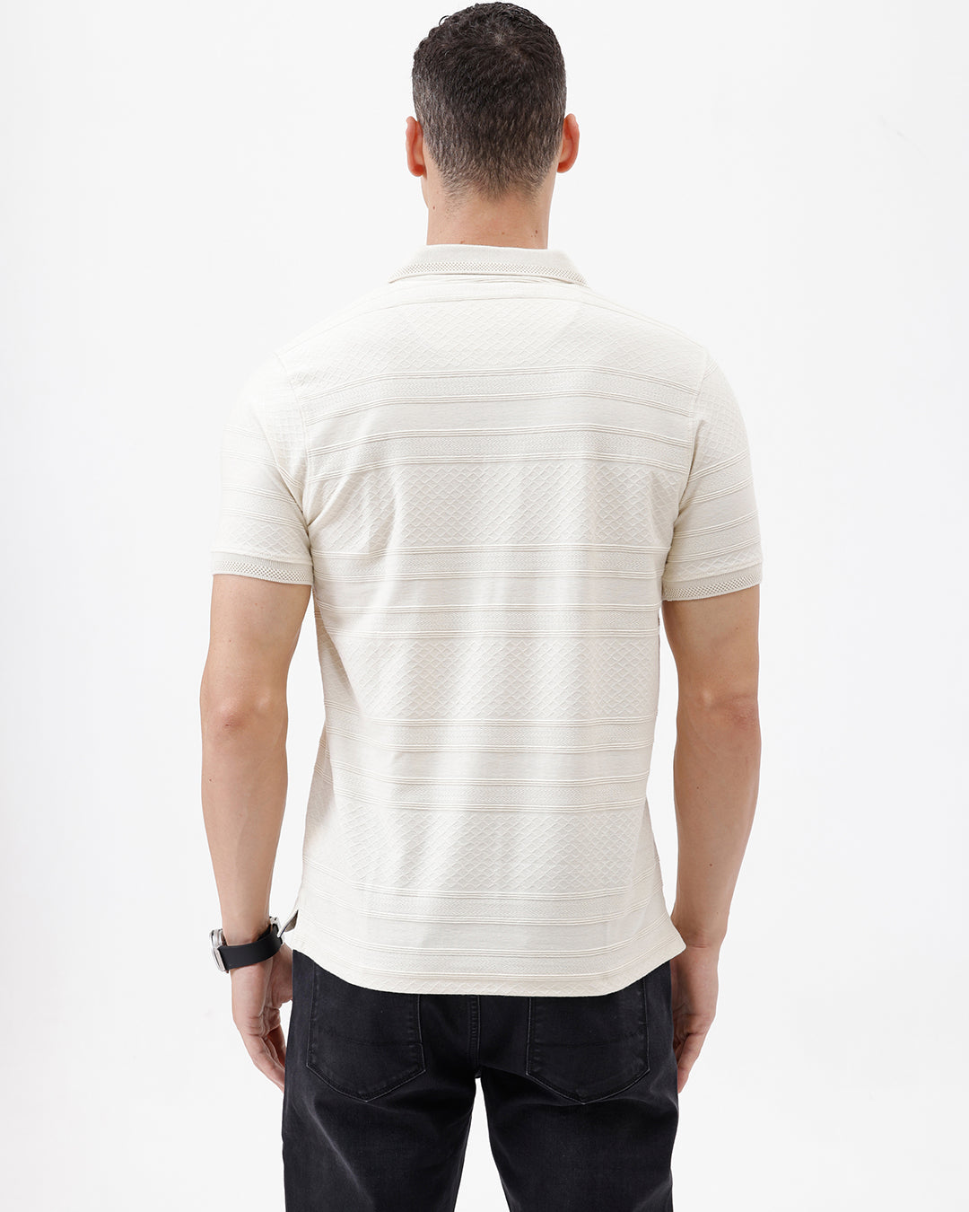 A man wearing a short-sleeve polo shirt in cream with subtle horizontal textured stripes, paired with dark jeans, standing against a plain white studio background.