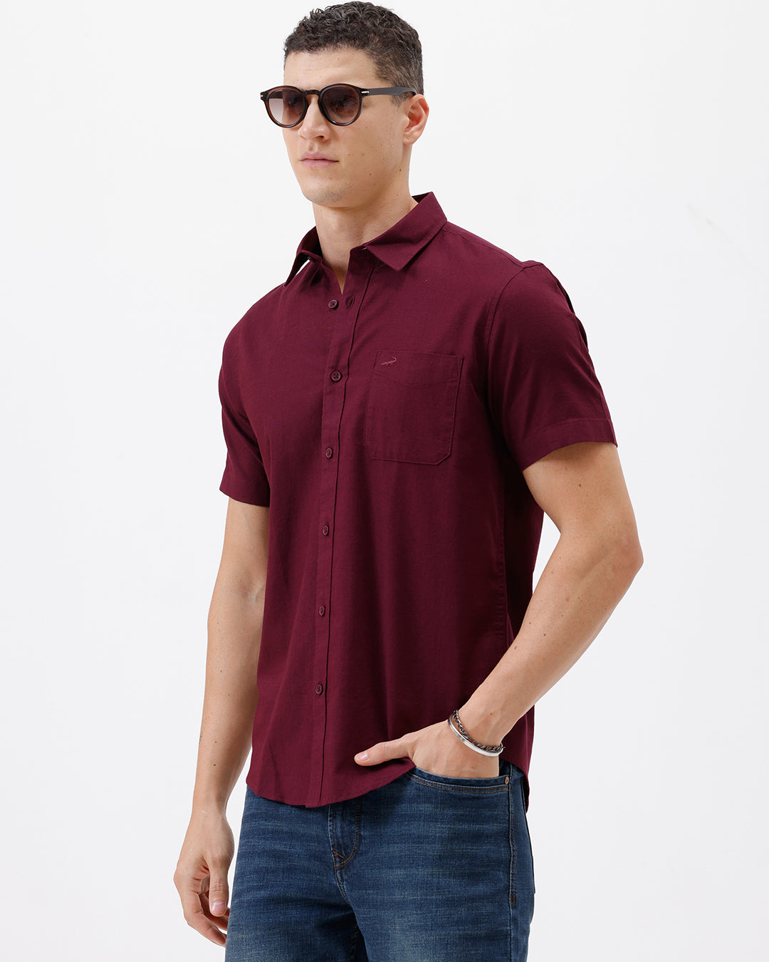 A man wearing a short-sleeve button-up shirt in maroon with a chest pocket, paired with blue jeans, standing against a plain white studio background.
