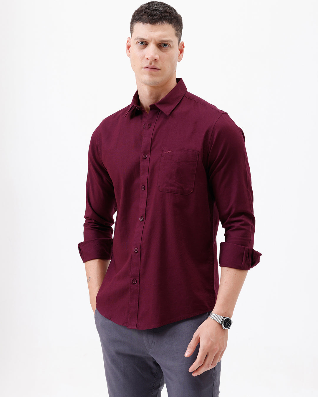A man wearing a long-sleeve button-down shirt in deep burgundy with rolled-up sleeves and gray trousers, standing against a plain white studio background.