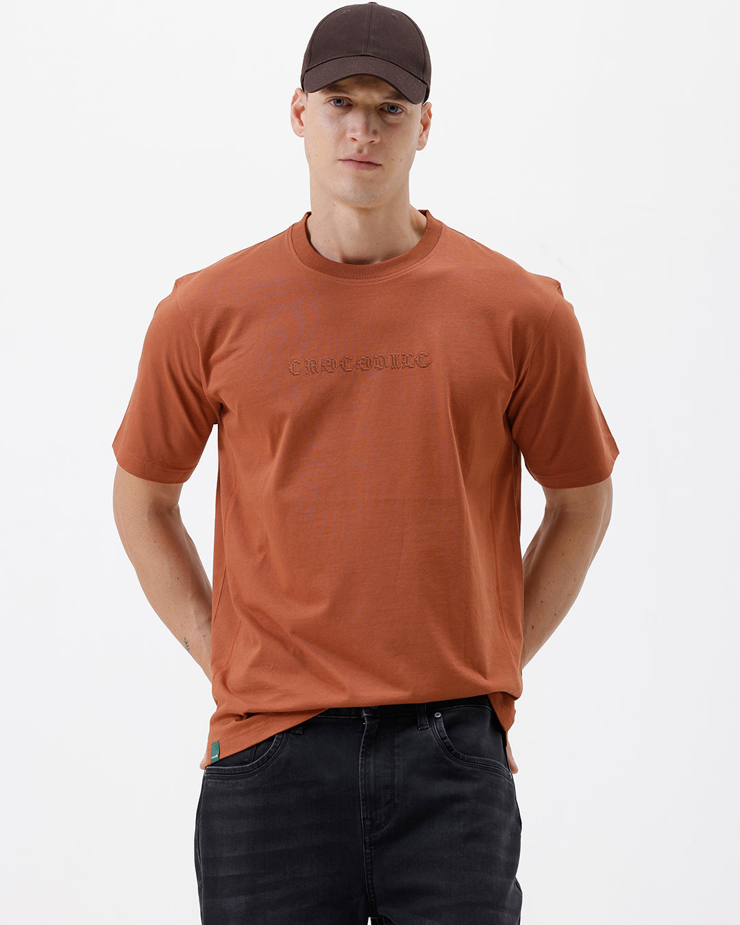 A man wearing a rust-orange crewneck t-shirt with a subtle tonal 'ELECTRIC' logo across the chest, paired with black pants, photographed against a clean white studio background.