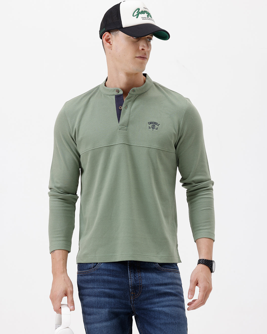 A man wearing a sage green long-sleeve henley shirt with a small embroidered logo on the left chest, paired with blue jeans, against a plain white studio background.