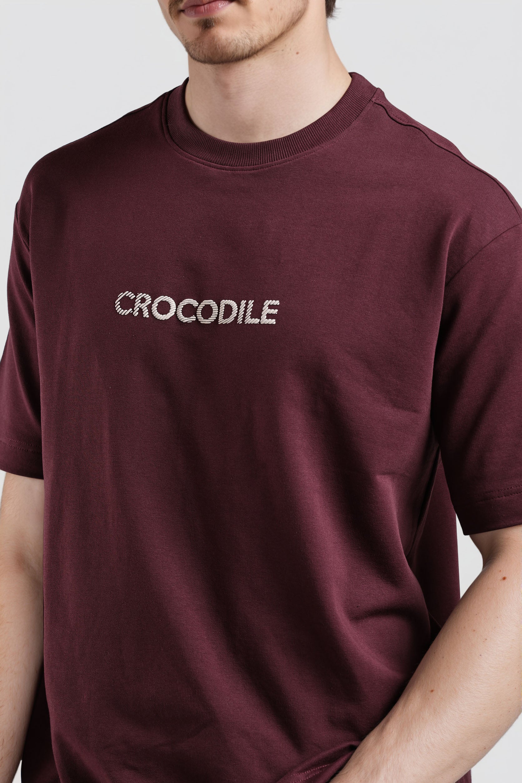 Maroon t-shirt with 'CROCODILE' logo worn by a person on a light gray background