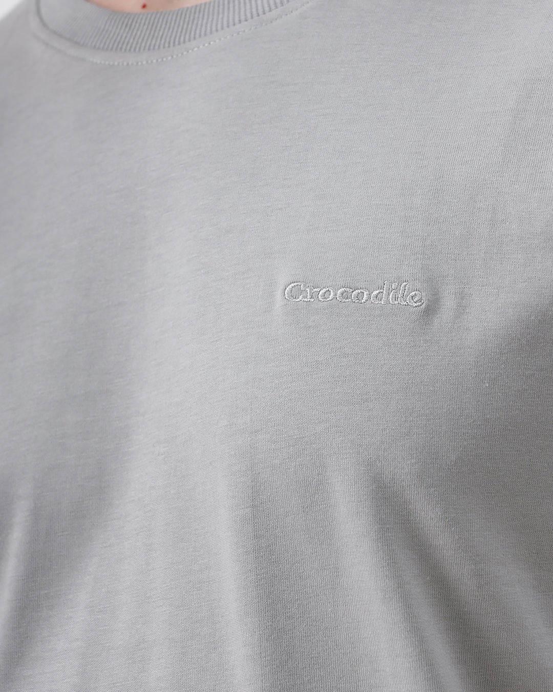A man wearing a light gray short-sleeve crewneck t-shirt with a subtle tonal chest logo, paired with dark jeans, standing against a light gray studio background.