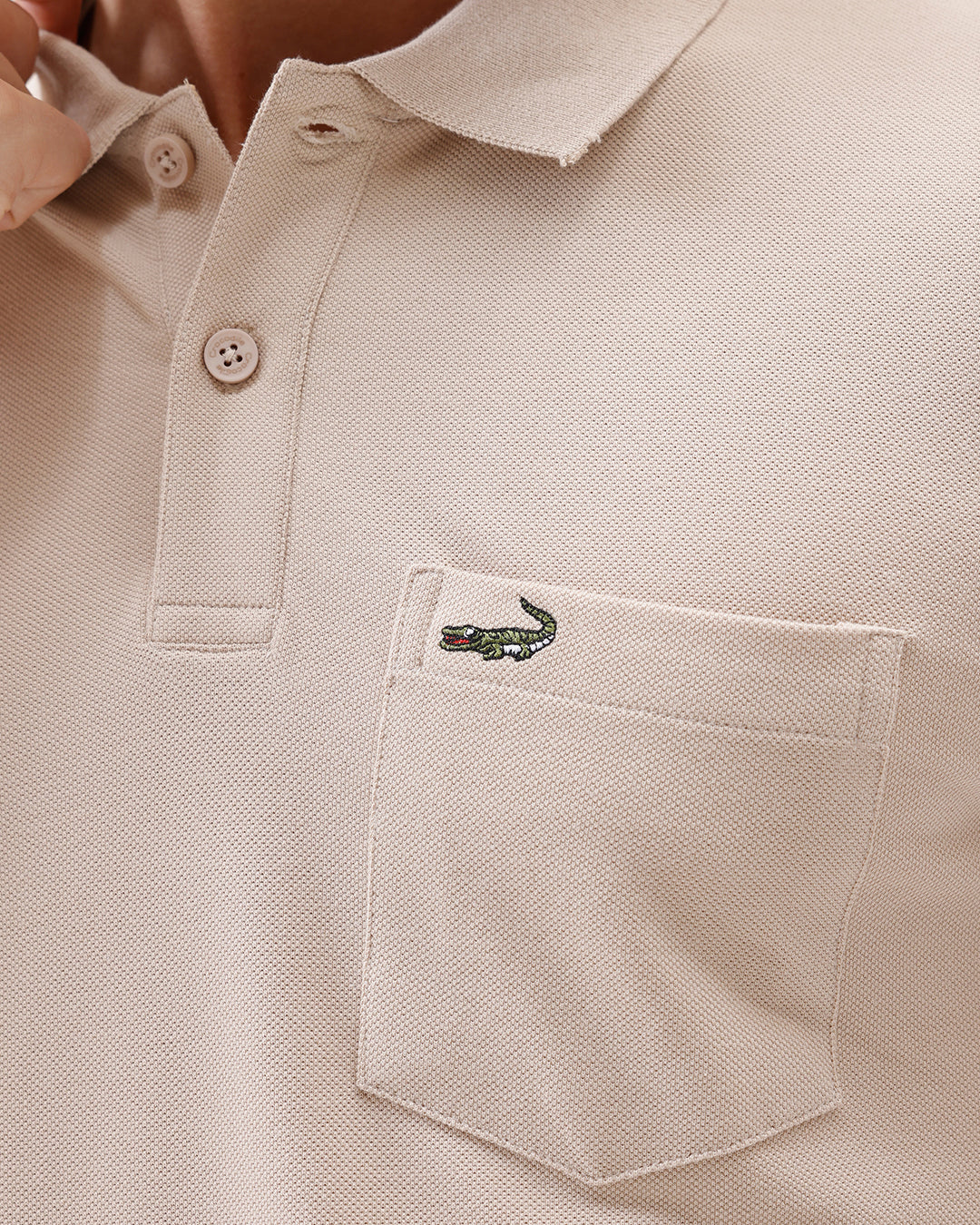 A man wearing a light beige short-sleeve polo shirt with a small crocodile logo on the chest pocket, paired with gray trousers, photographed against a plain white studio background.