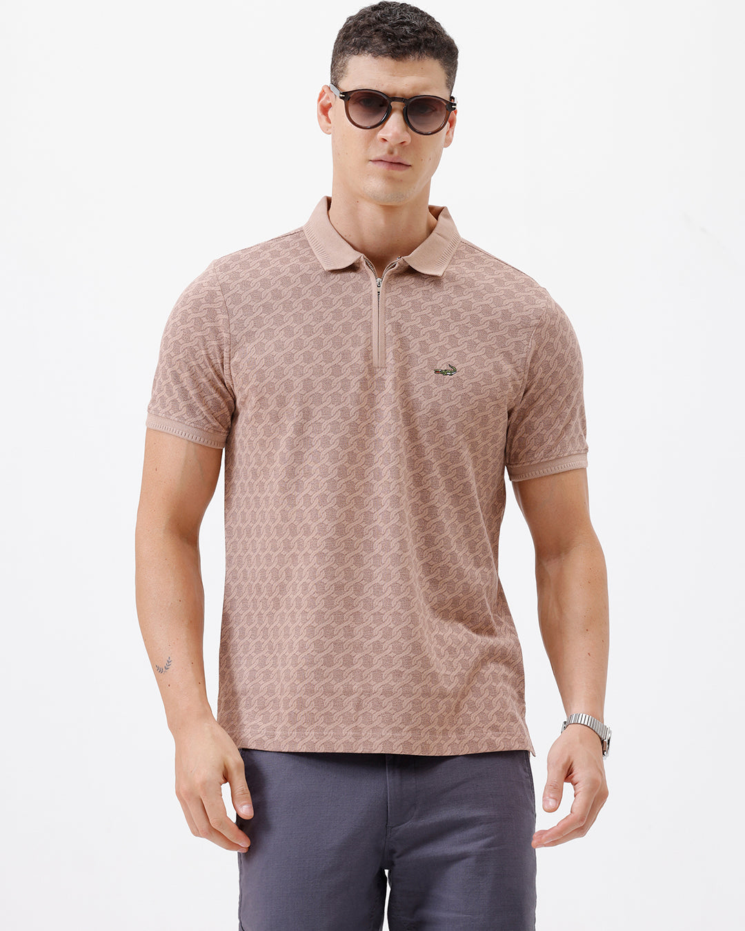 A man wearing a short-sleeve polo shirt with a muted brown and beige geometric pattern, paired with dark gray trousers, standing against a plain white studio background.