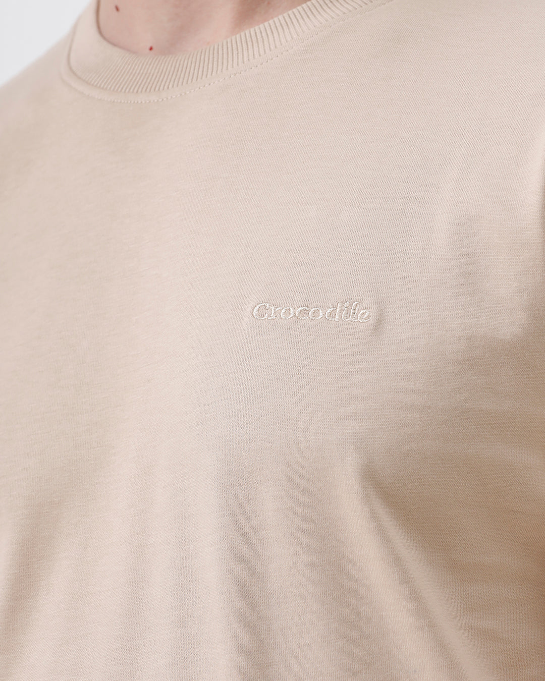 A man wearing a beige short-sleeve crewneck t-shirt with a subtle tonal logo on the left chest, paired with black jeans, standing against a light gray studio background.