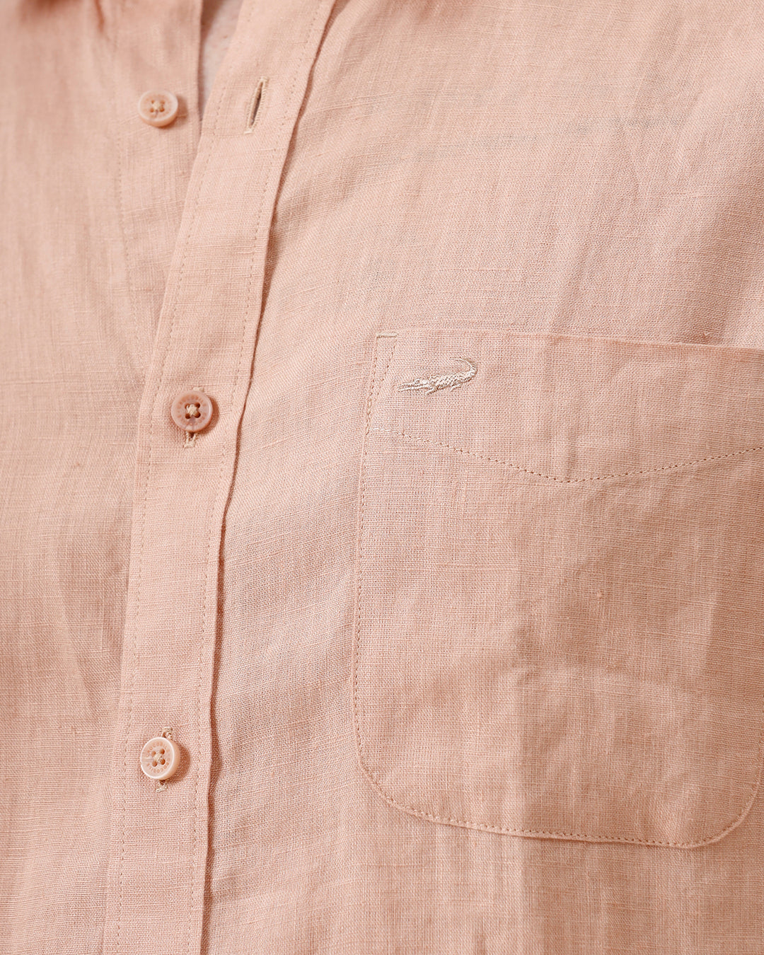 A man wearing a short-sleeve linen button-up shirt in a light peach-pink color, paired with light gray trousers, standing against a plain light gray background.