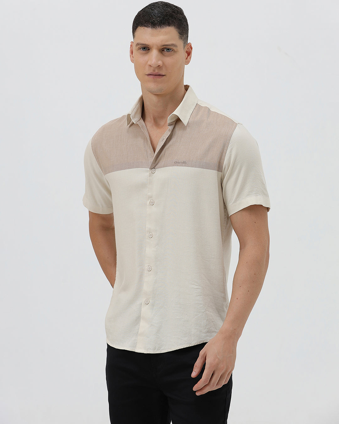 A man wearing a short-sleeve linen button-up shirt with a color-blocked design in beige and cream, paired with black pants, against a plain white studio background.