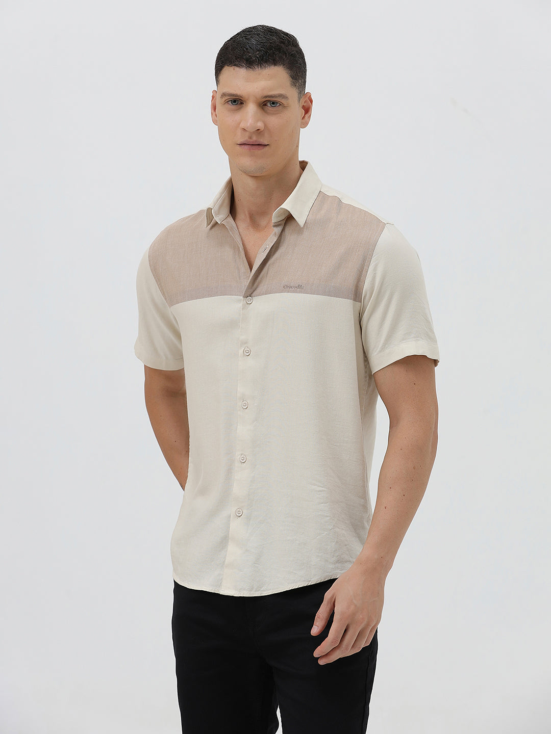 A man wearing a short-sleeve linen button-up shirt with a color-blocked design in beige and cream, paired with black pants, against a plain white studio background.