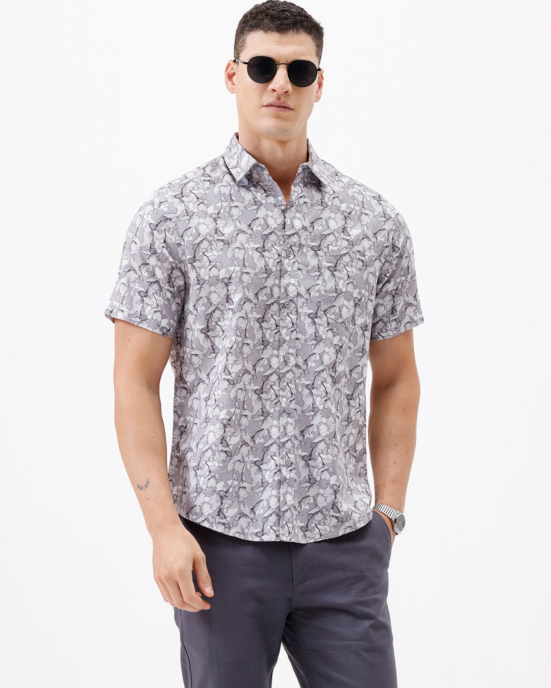 A man wearing a short-sleeve button-up shirt with a gray and white abstract pattern, paired with dark gray trousers, standing against a plain white studio background.