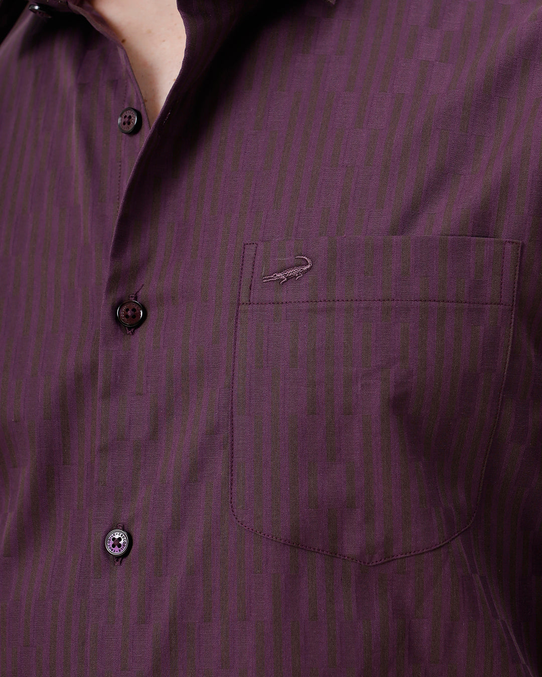 A man wearing a dark purple long-sleeve button-down shirt with subtle vertical stripes and dark navy trousers, standing against a light gray background.