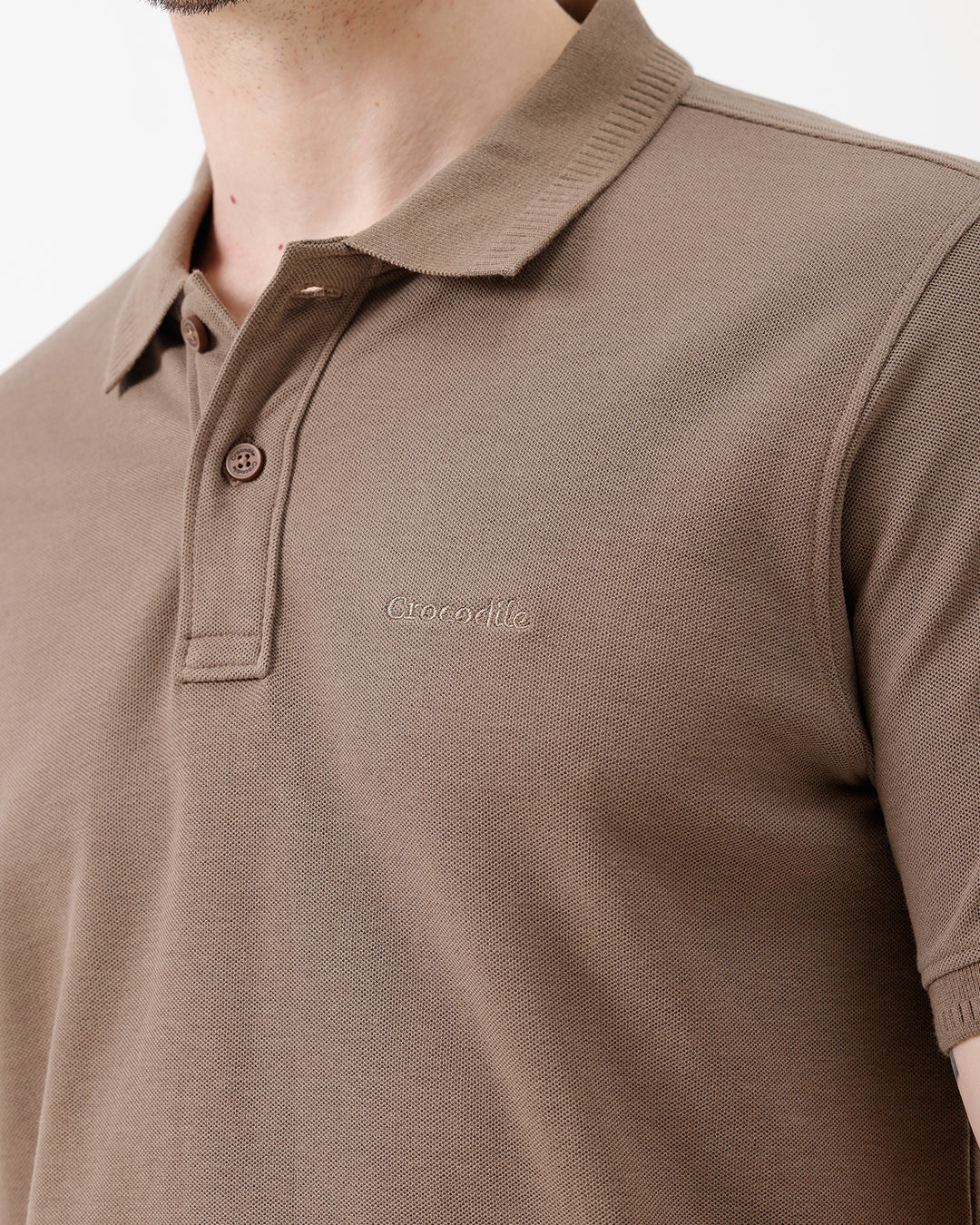 A man wearing a brown short-sleeve polo shirt with dark navy pants, standing against a plain light gray studio background.