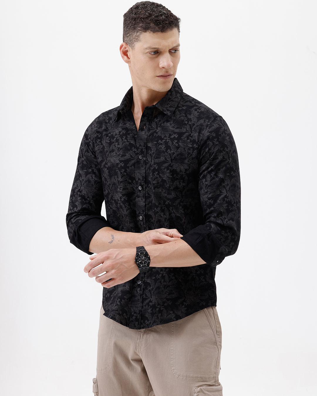 A man wearing a black long-sleeve button-up shirt with a subtle tonal floral pattern, paired with beige cargo pants and dark sunglasses, standing against a plain white background.