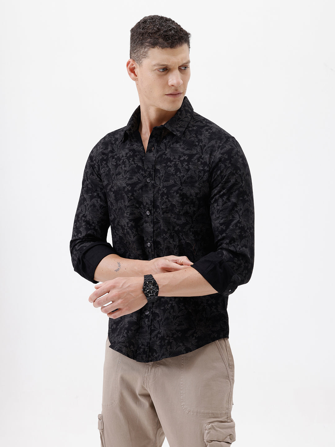 A man wearing a black long-sleeve button-up shirt with a subtle tonal floral pattern, paired with beige cargo pants and dark sunglasses, standing against a plain white background.