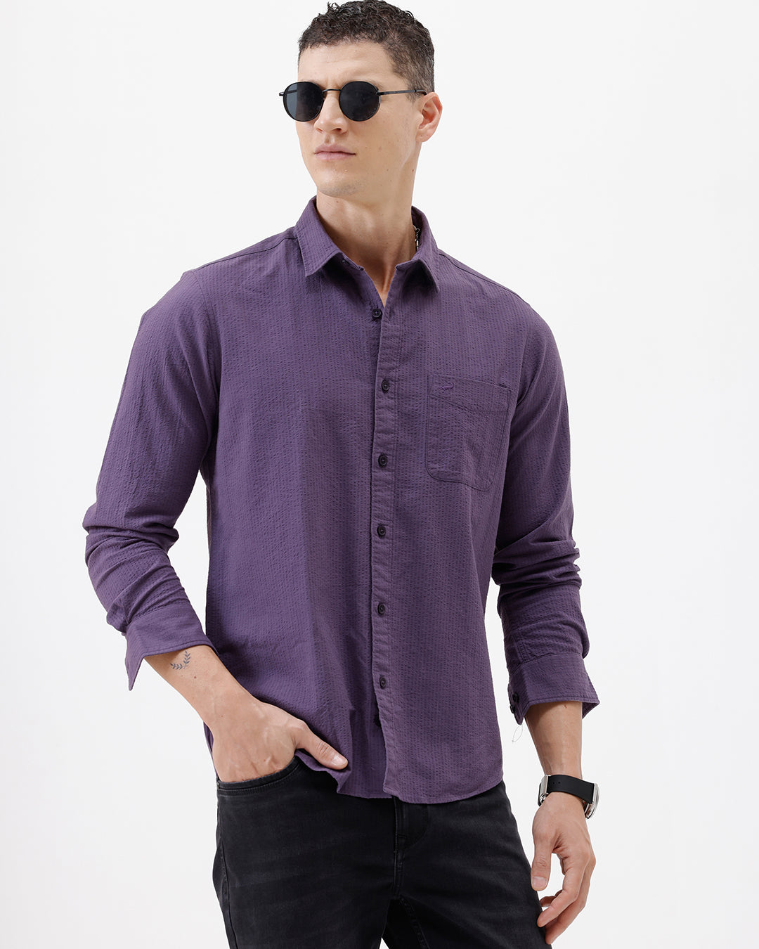 A man wearing a long-sleeve button-down shirt in a muted purple color, paired with black pants, standing against a plain white studio background.
