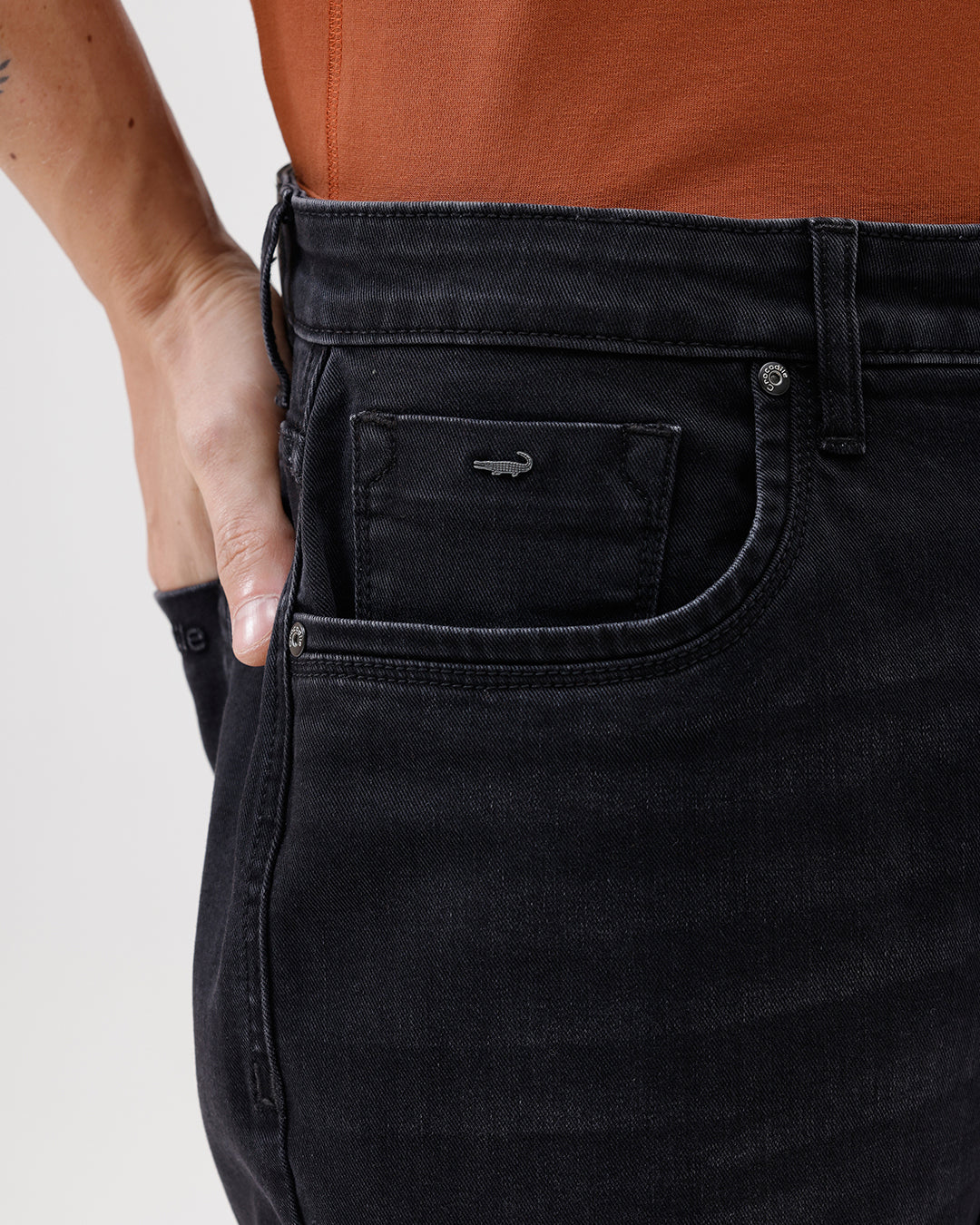 A person wearing black slim-fit jeans, paired with a rust-orange t-shirt and brown sneakers, shown from the waist down against a plain white studio background.