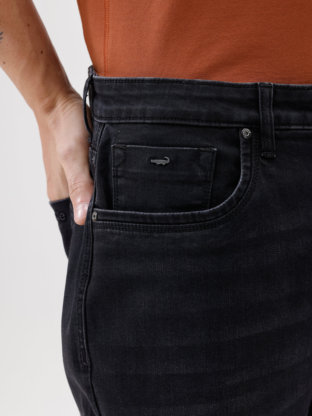 A person wearing black slim-fit jeans, paired with a rust-orange t-shirt and brown sneakers, shown from the waist down against a plain white studio background.