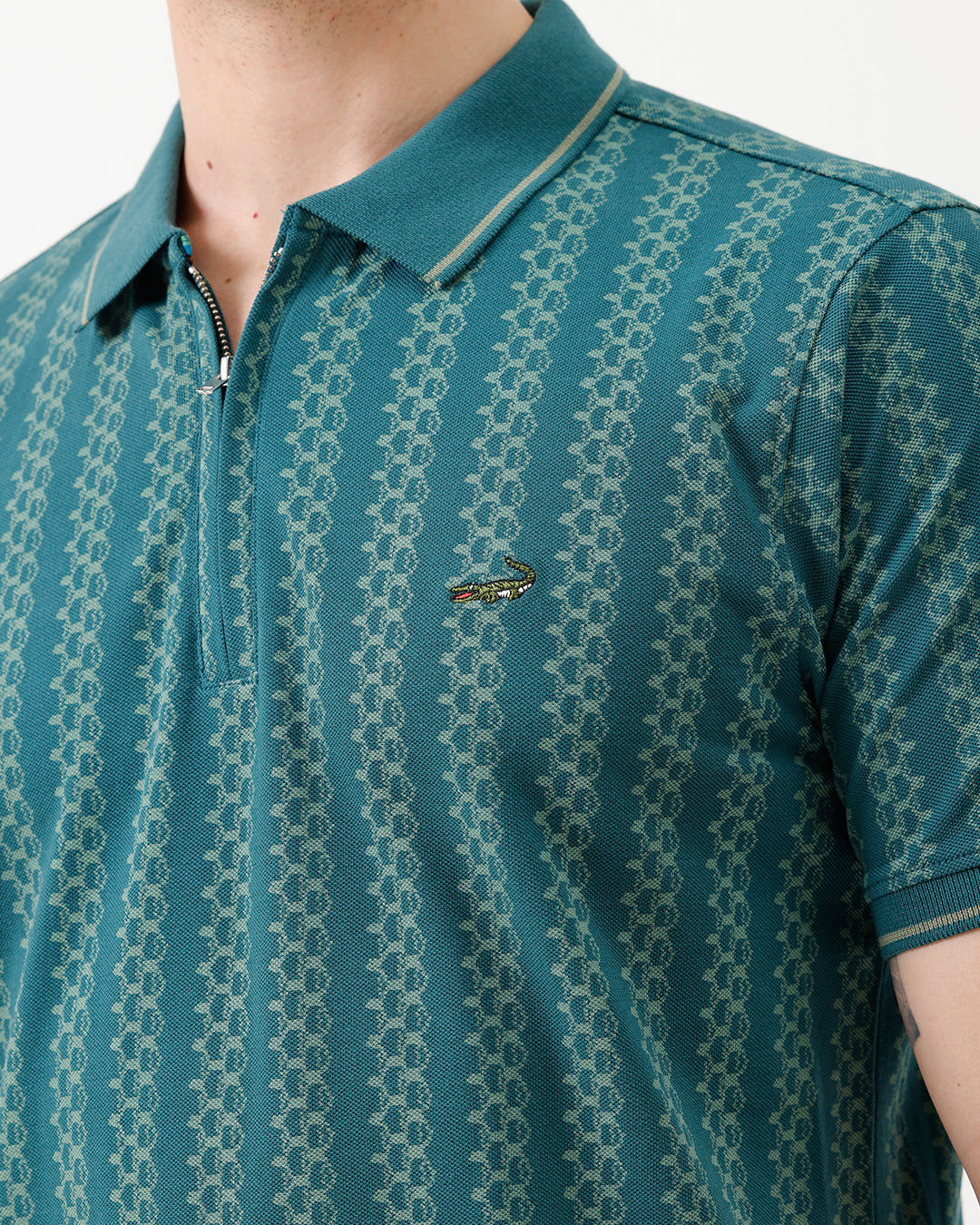 A man wearing a teal green short-sleeve polo shirt with a white geometric pattern, paired with black jeans, standing against a plain white studio background.