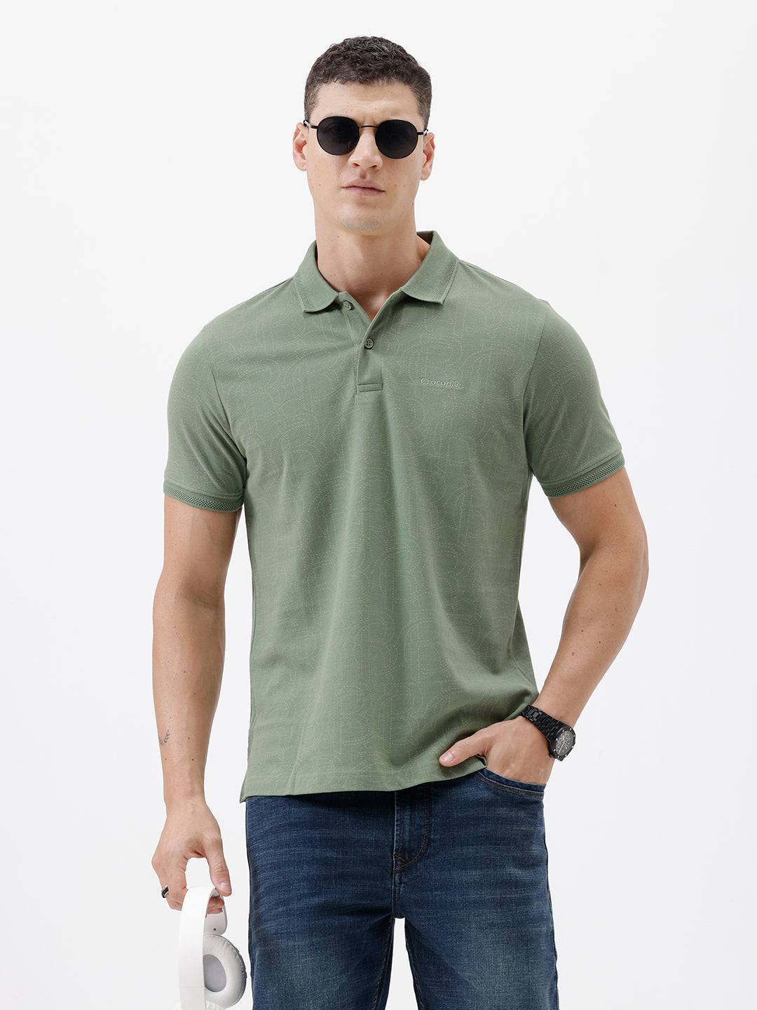 A man wearing a short-sleeve polo shirt in sage green, paired with dark blue jeans, standing against a plain light gray background.