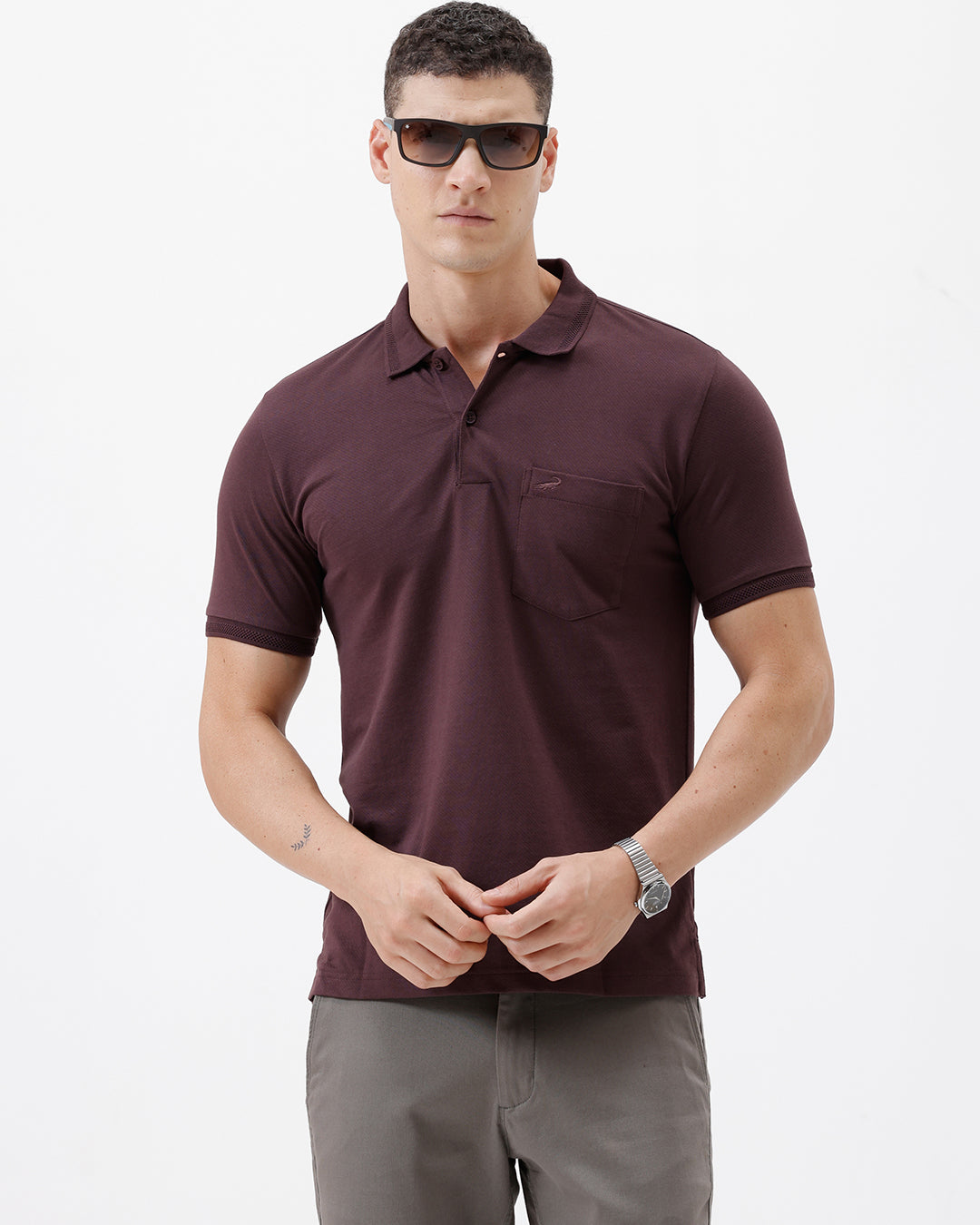 A man wearing a short-sleeve polo shirt in dark burgundy with a chest pocket, paired with gray trousers, standing against a plain light gray background.