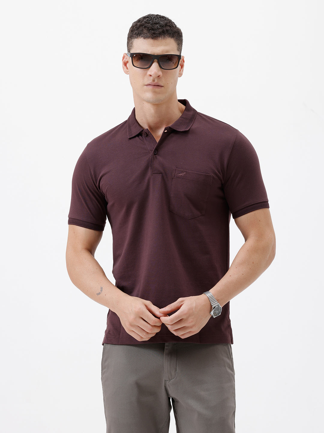 A man wearing a short-sleeve polo shirt in dark burgundy with a chest pocket, paired with gray trousers, standing against a plain light gray background.
