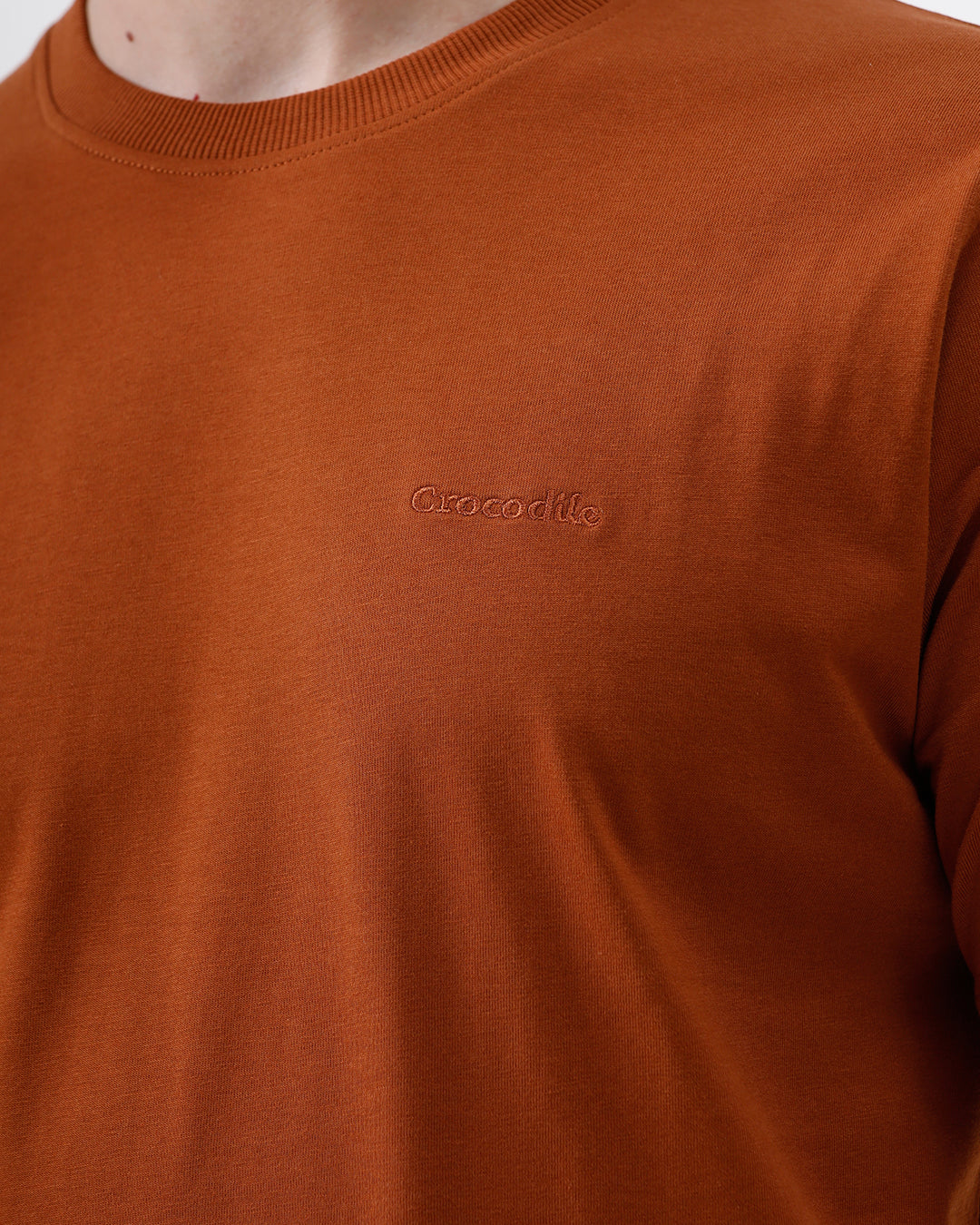 A man wearing a rust-orange crew-neck t-shirt and dark blue jeans, standing against a light gray studio background.