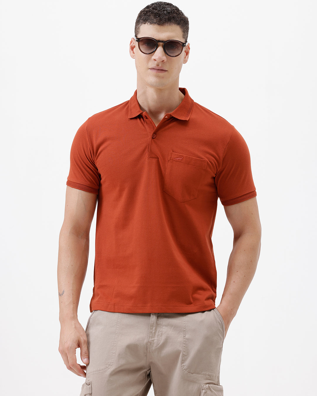 A man wearing a rust-orange short-sleeve polo shirt with a chest pocket, paired with beige cargo pants, standing against a plain light gray studio background.