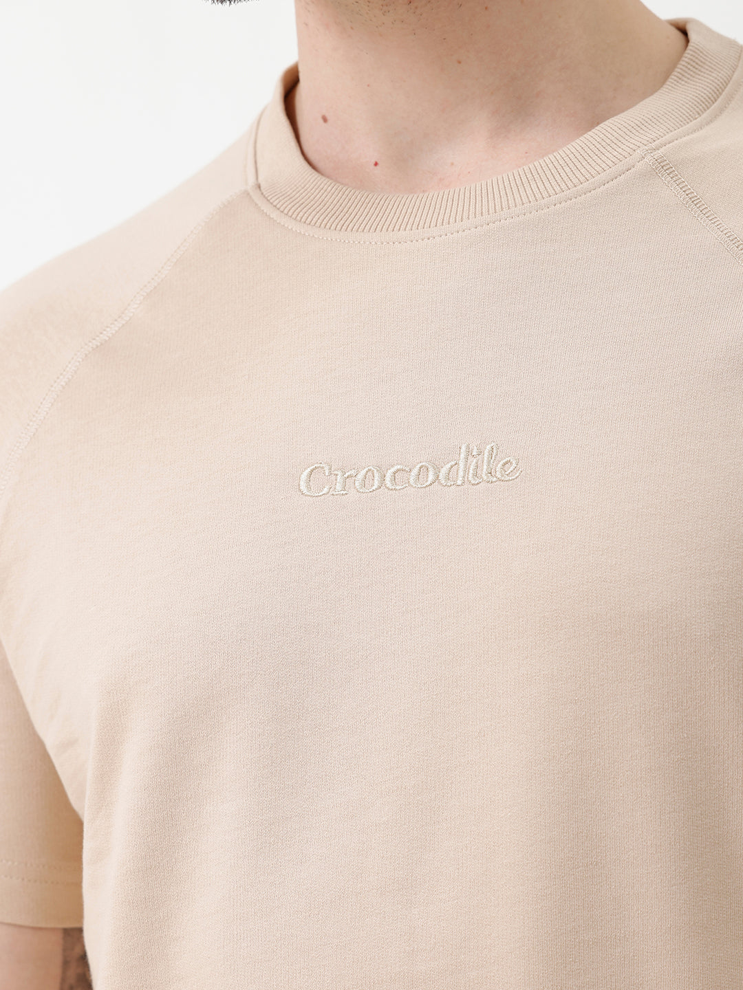 A man wearing a light beige short-sleeve t-shirt with a subtle tonal logo on the chest, paired with black pants, standing against a plain white studio background.