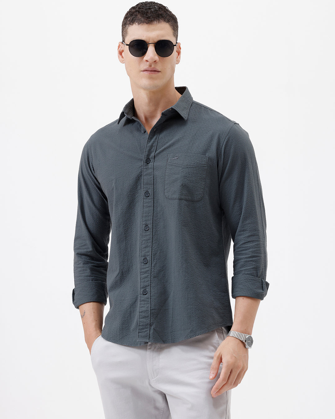 A man wearing a dark charcoal gray long-sleeve button-down shirt with a chest pocket, paired with light gray trousers, standing against a plain white studio background.