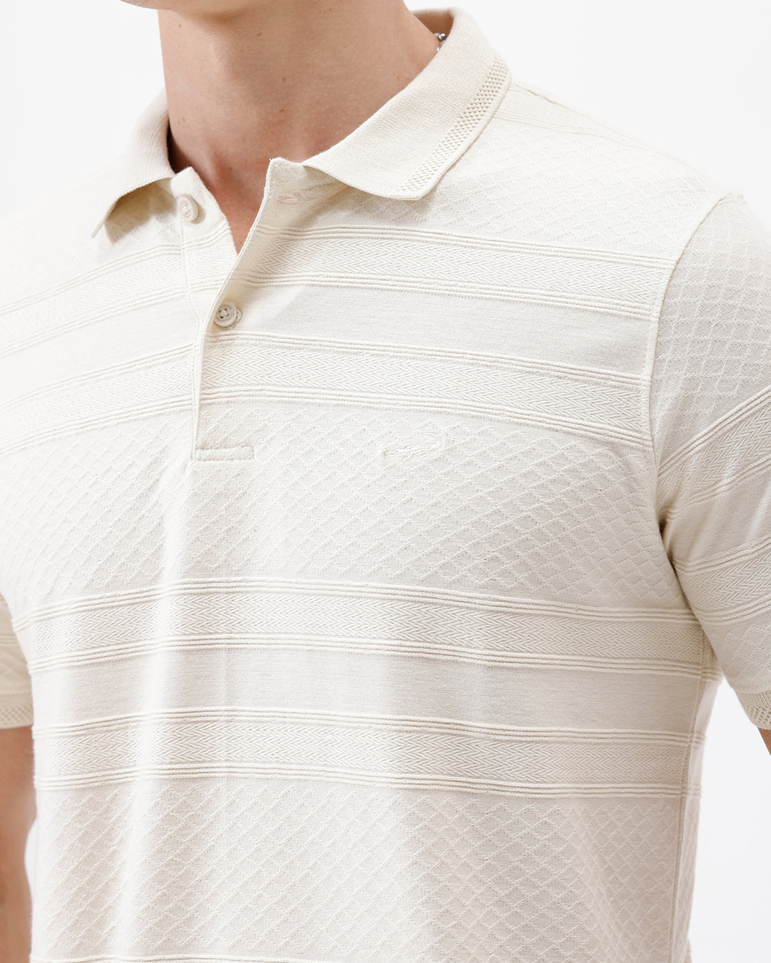 A man wearing a short-sleeve polo shirt in cream with subtle horizontal textured stripes, paired with dark jeans, standing against a plain white studio background.