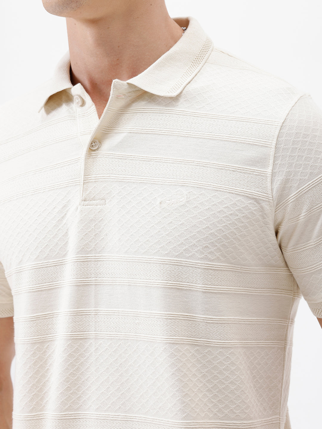 A man wearing a short-sleeve polo shirt in cream with subtle horizontal textured stripes, paired with dark jeans, standing against a plain white studio background.