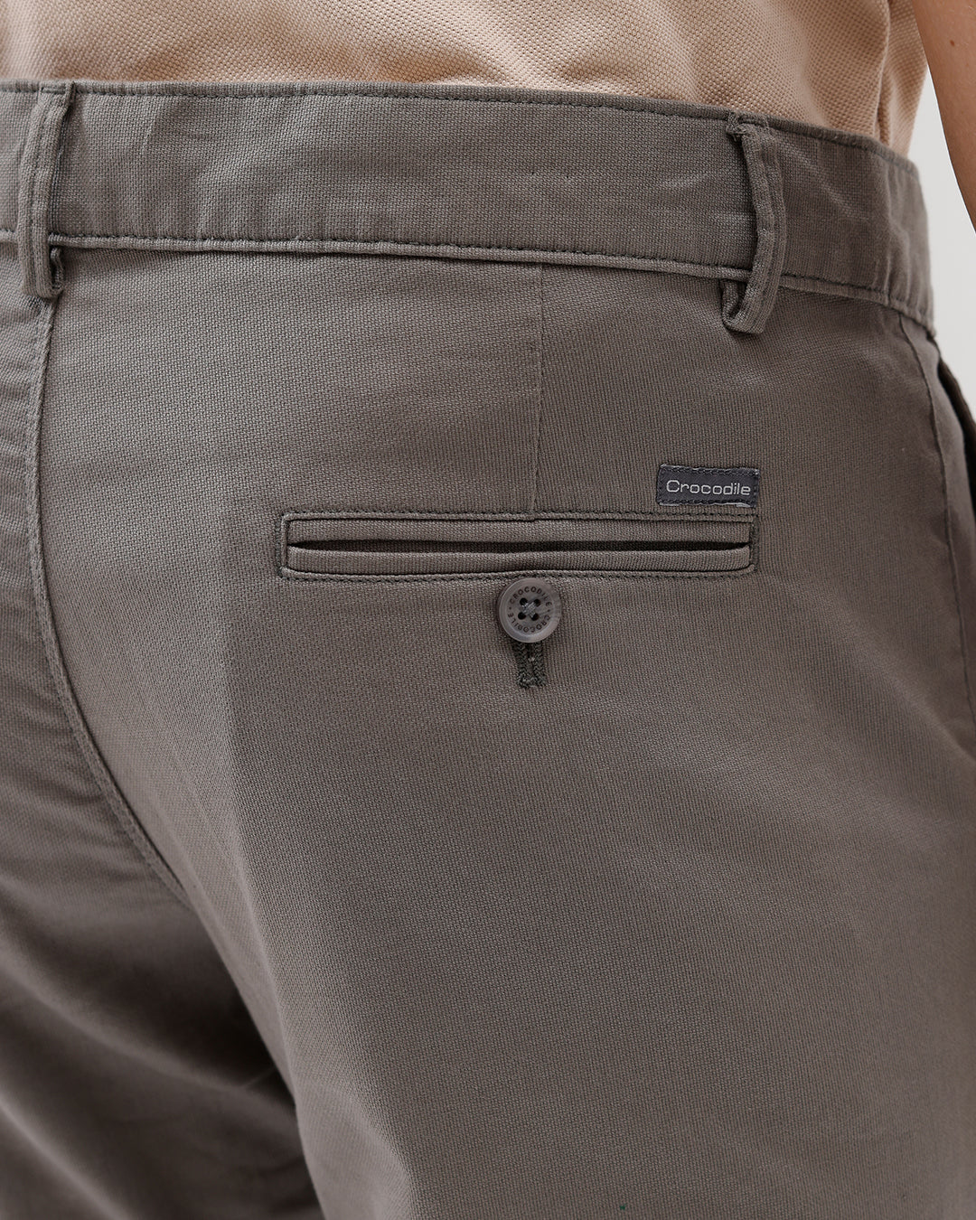 A man wearing slim-fit taupe gray chino pants, paired with a beige t-shirt and brown sneakers, shown from the waist down on a white studio background.