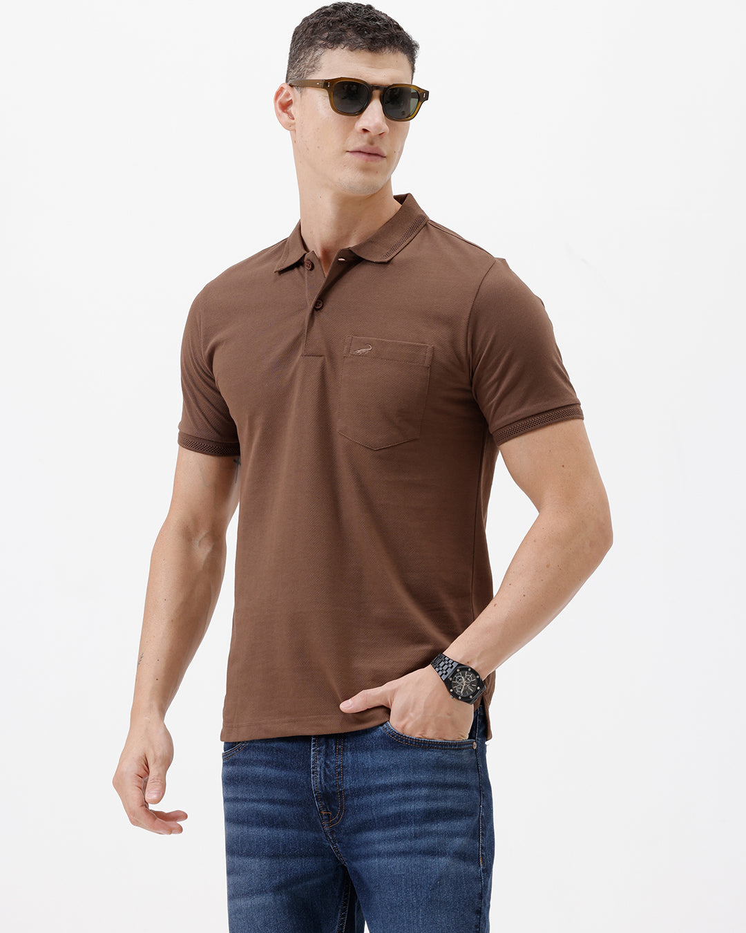A man wearing a brown short-sleeve polo shirt with a chest pocket, paired with blue jeans, standing against a plain light gray background.