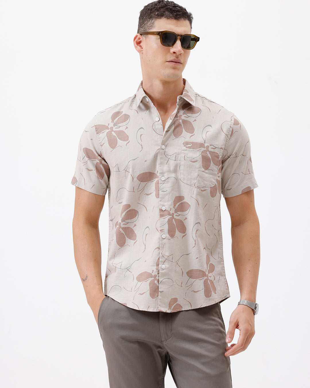 A man wearing a short-sleeve button-up shirt with a beige and brown floral pattern, paired with gray trousers, standing against a plain white background.
