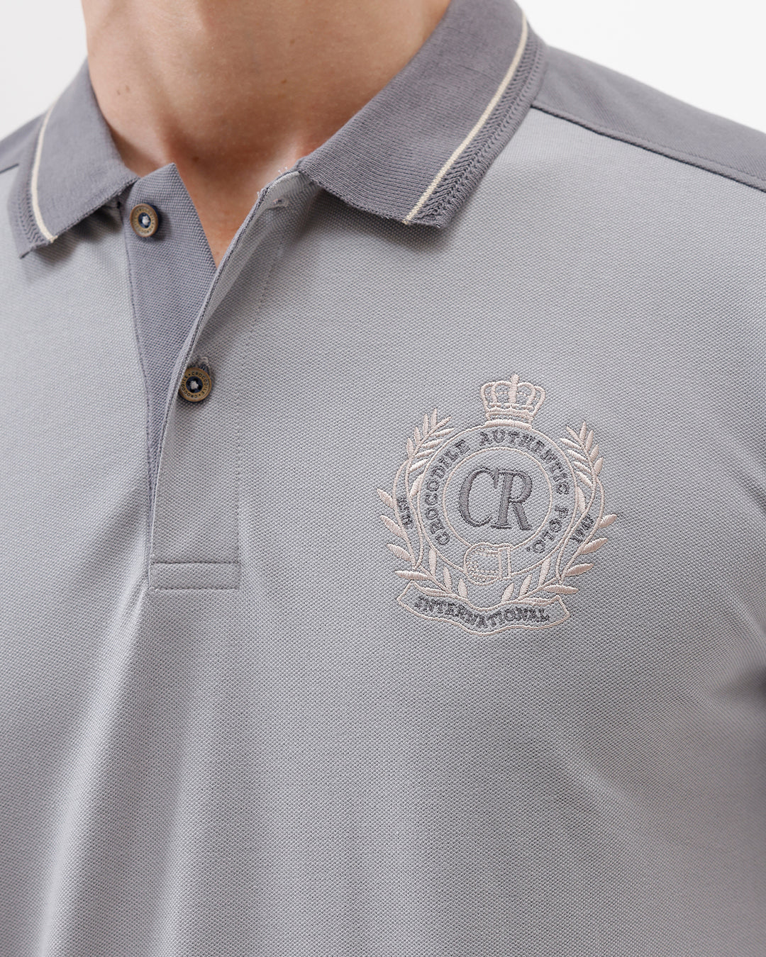 A man wearing a light gray short-sleeve polo shirt with darker gray shoulder panels and a small crest logo on the left chest, paired with khaki cargo pants, against a plain light gray studio background.
