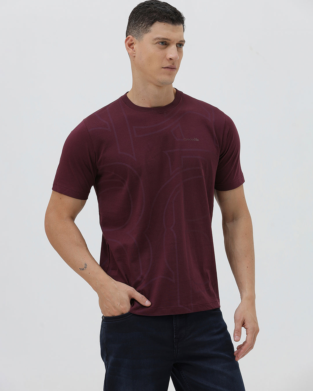 A man wearing a short-sleeve crewneck t-shirt in maroon with a subtle tonal graphic pattern, paired with dark blue jeans, against a light gray studio background.