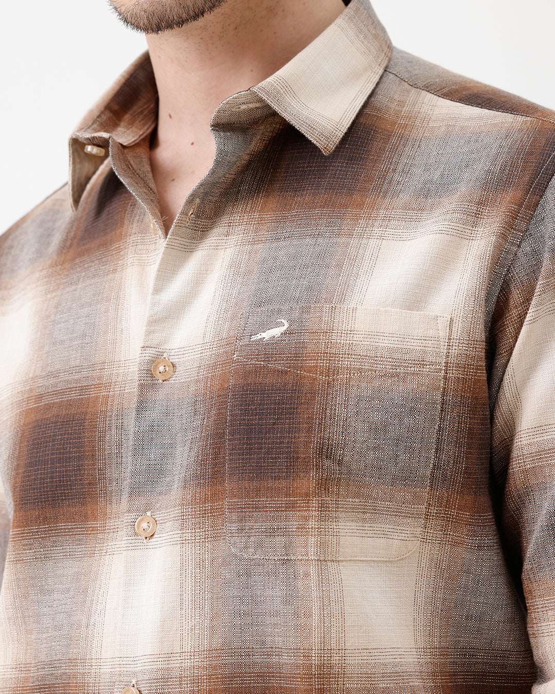 A man wearing a long-sleeve button-up plaid shirt in brown, beige, and gray tones, paired with dark jeans, standing against a plain white studio background.