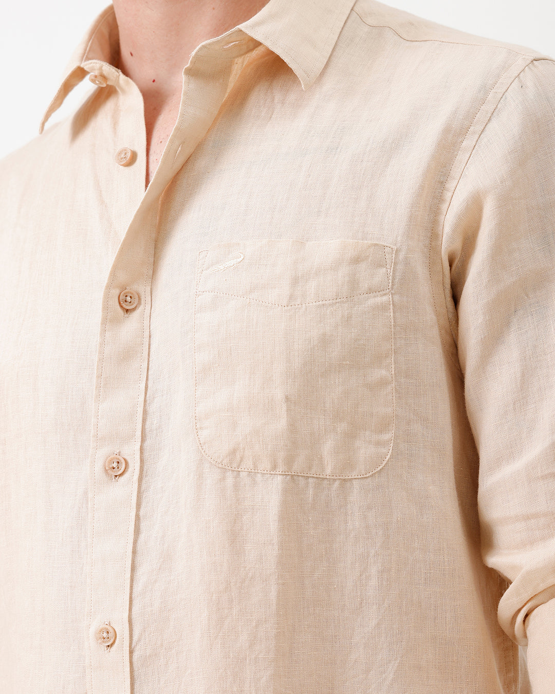 A man wearing a light beige linen button-up shirt with rolled-up sleeves and dark jeans, standing against a plain white studio background.