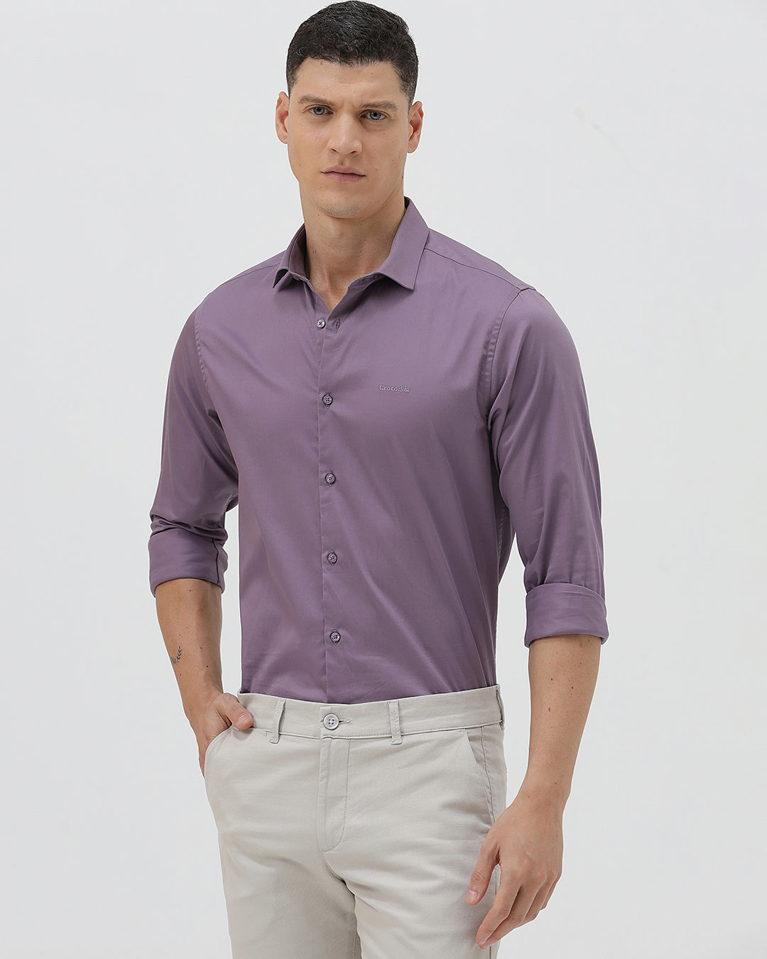 A man wearing a purple long-sleeve button-down shirt with rolled-up sleeves and light beige trousers, standing against a plain light gray background.