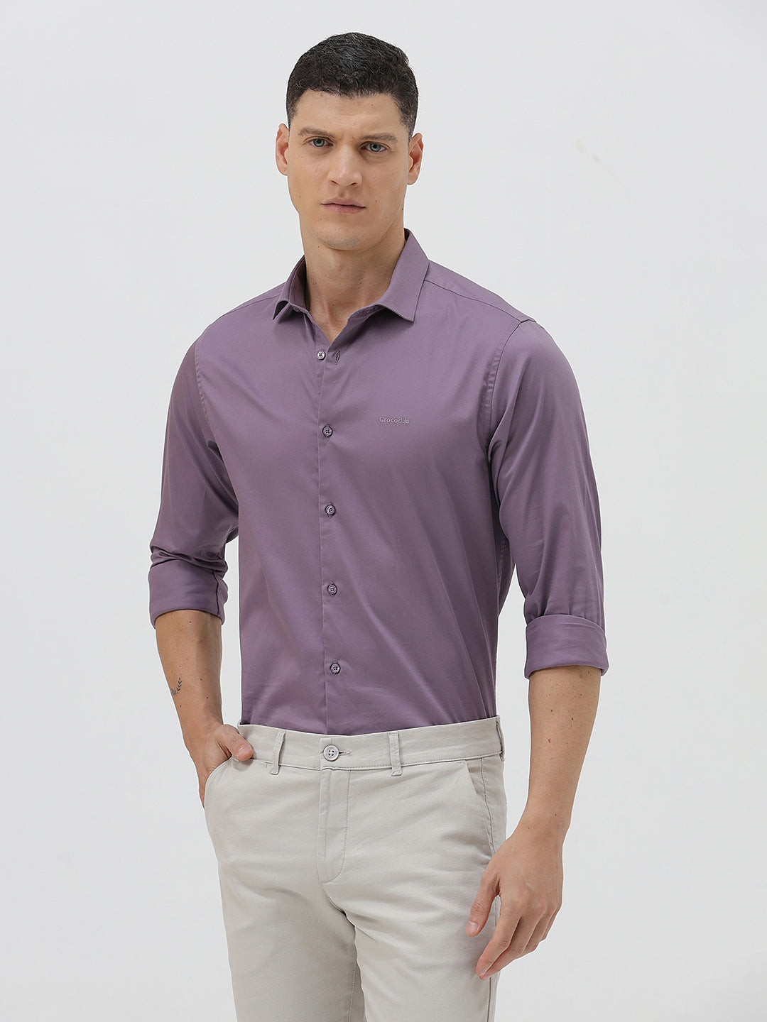 A man wearing a purple long-sleeve button-down shirt with rolled-up sleeves and light beige trousers, standing against a plain light gray background.