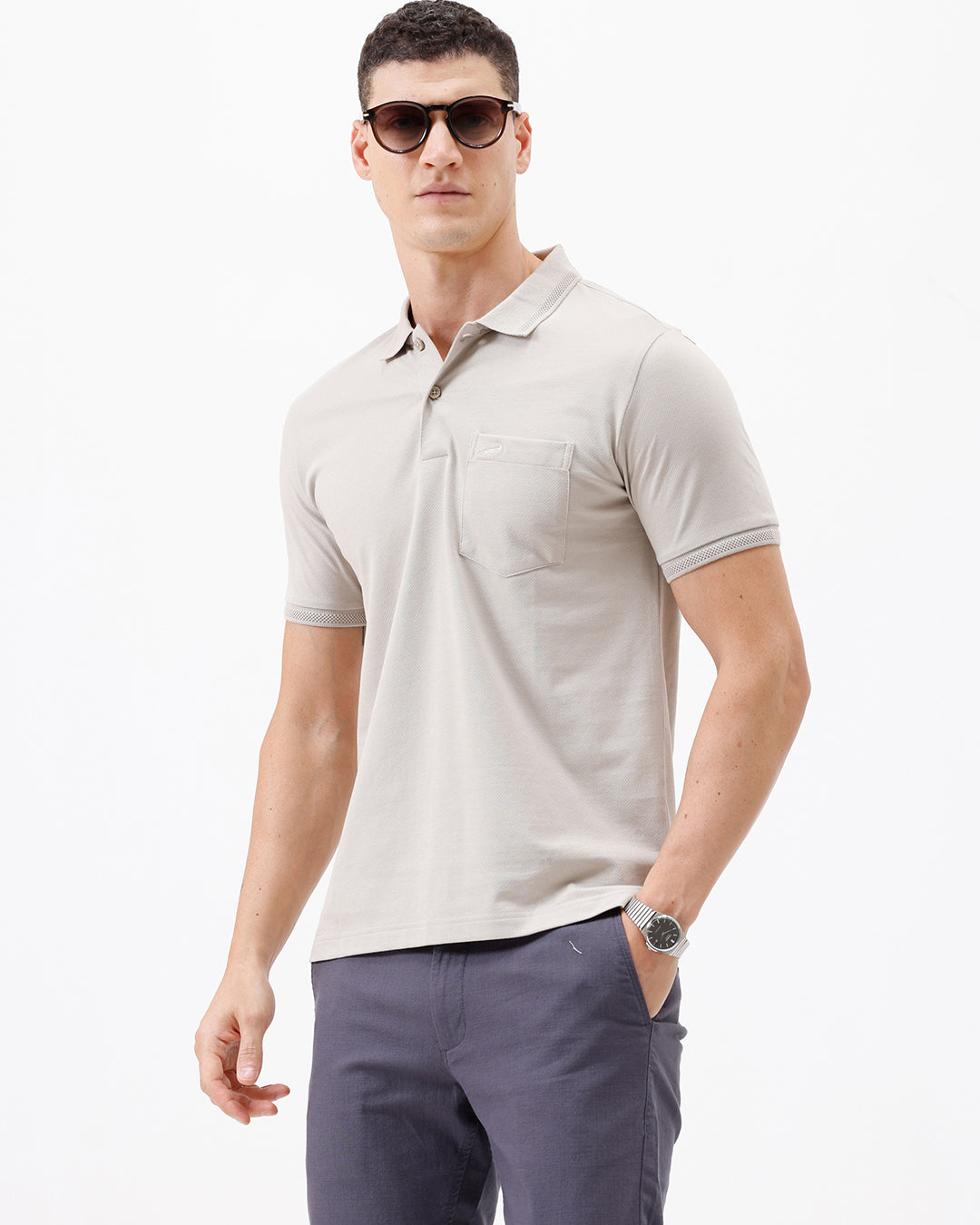 A man wearing a light beige short-sleeve polo shirt with a chest pocket, paired with dark gray trousers, standing against a plain white studio background.