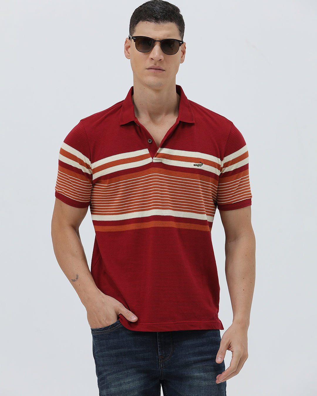 A man wearing a red short-sleeve polo shirt with horizontal cream and rust-orange stripes across the chest and sleeves, paired with dark blue jeans and black sunglasses, against a light gray studio background.