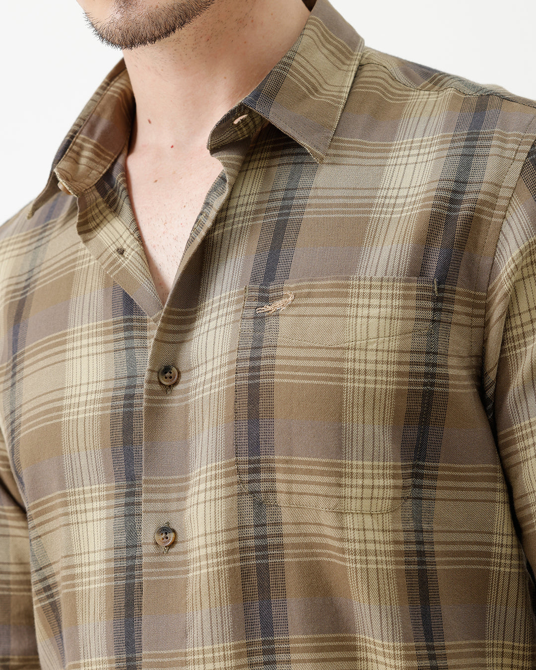 A man wearing a brown and beige plaid long-sleeve button-down shirt with dark sunglasses and black jeans, standing against a plain white studio background.