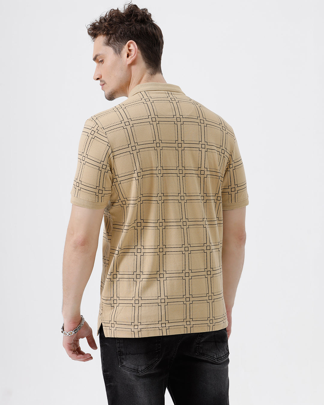 A man wearing a beige short-sleeve polo shirt with a brown grid pattern, paired with black jeans, standing against a plain white studio background.