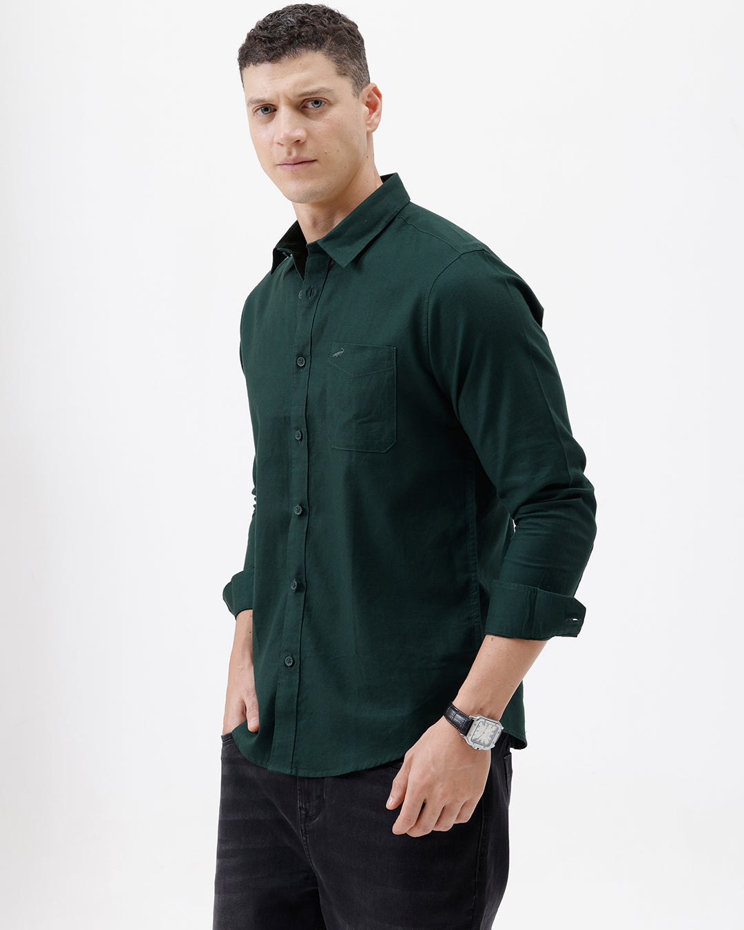A man wearing a dark forest green long-sleeve button-down shirt with a chest pocket, paired with dark jeans, standing against a plain white studio background.
