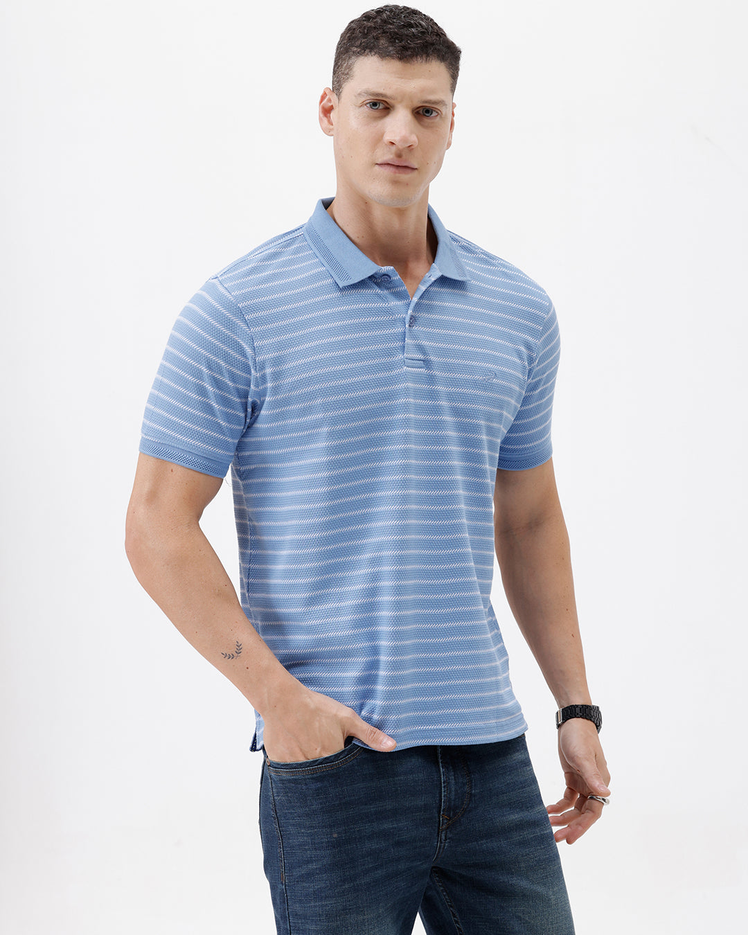 A man wearing a light blue and white striped short-sleeve polo shirt with dark blue jeans, standing against a plain light gray background.