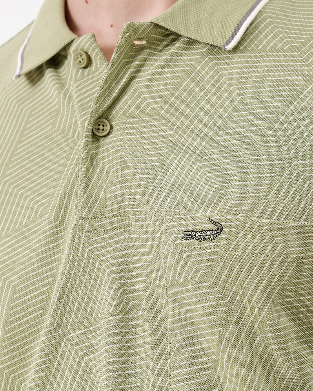 A man wearing a light sage green short-sleeve polo shirt with a subtle geometric pattern, paired with beige trousers, standing against a plain white studio background.